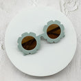 Load image into Gallery viewer, Round sunglasses with light sage green flower-shaped frames and brown lenses, displayed on a white circular surface.