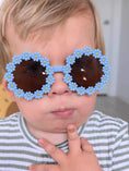 Load image into Gallery viewer, A toddler wearing adorable blue flower-shaped sunglasses with dark lenses, striking a playful pose with two fingers near their mouth while wearing a striped shirt.
