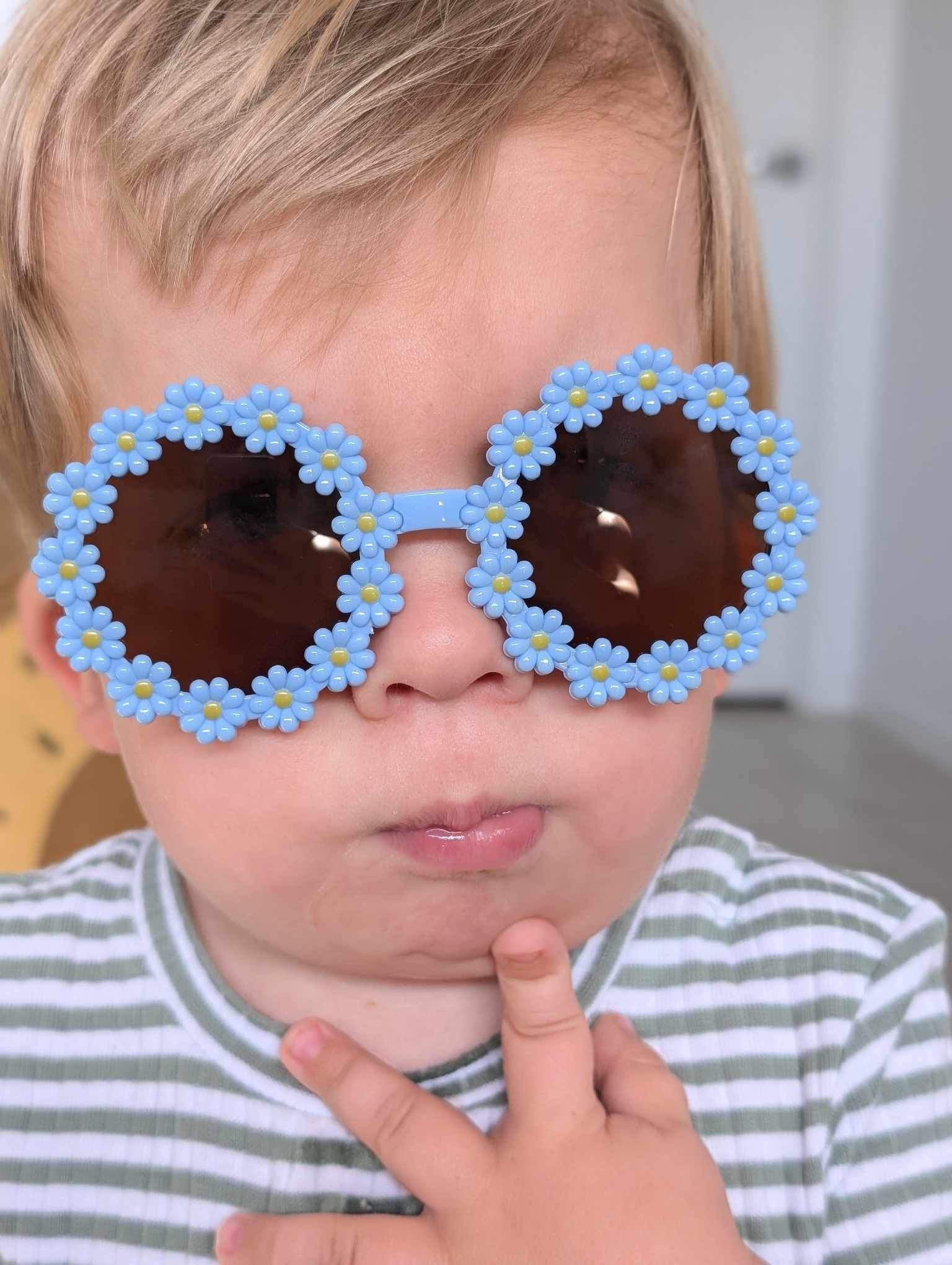 A toddler wearing adorable blue flower-shaped sunglasses with dark lenses, striking a playful pose with two fingers near their mouth while wearing a striped shirt.