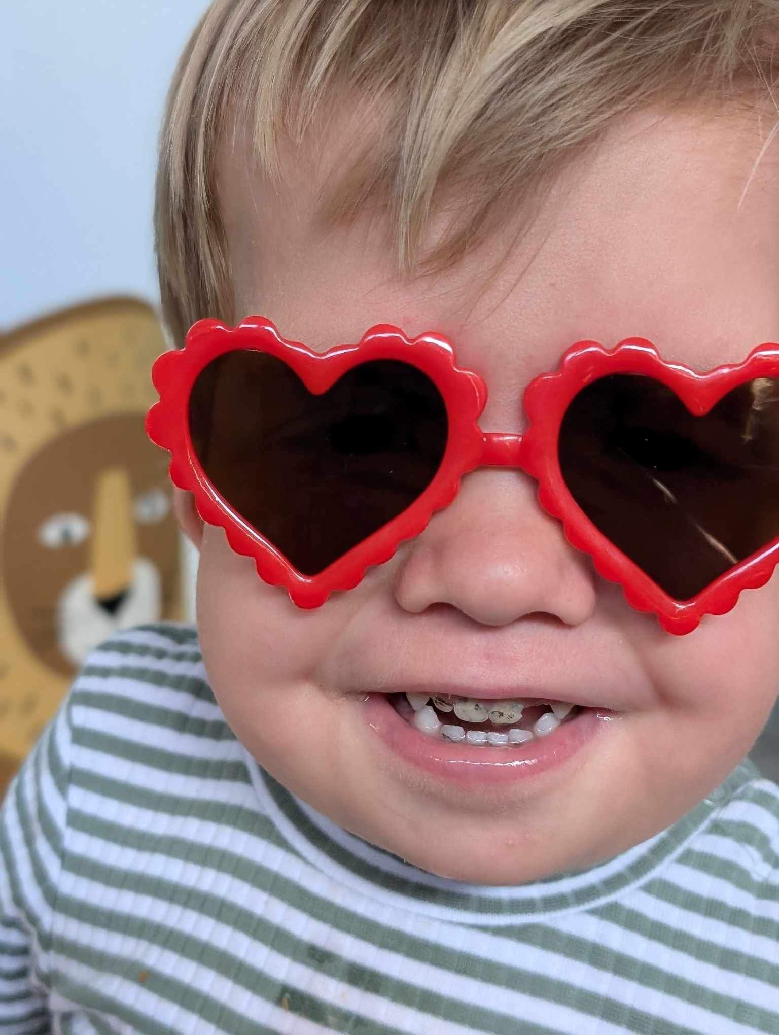 Smiling toddler wearing red heart-shaped sunglasses and a striped shirt.