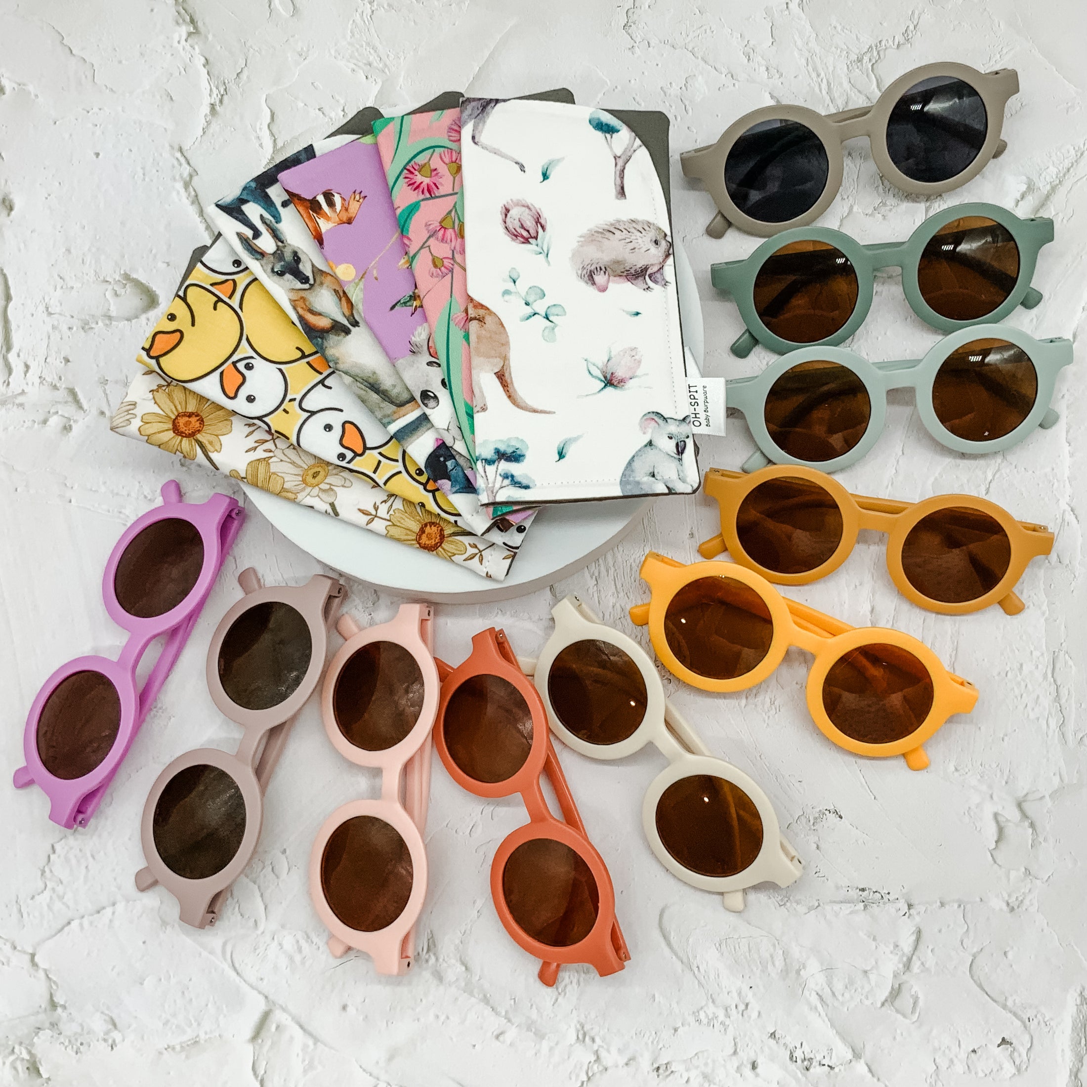 A colourful assortment of baby sunglasses and patterned fabric bibs displayed on a white textured surface, showcasing playful designs and vibrant hues.