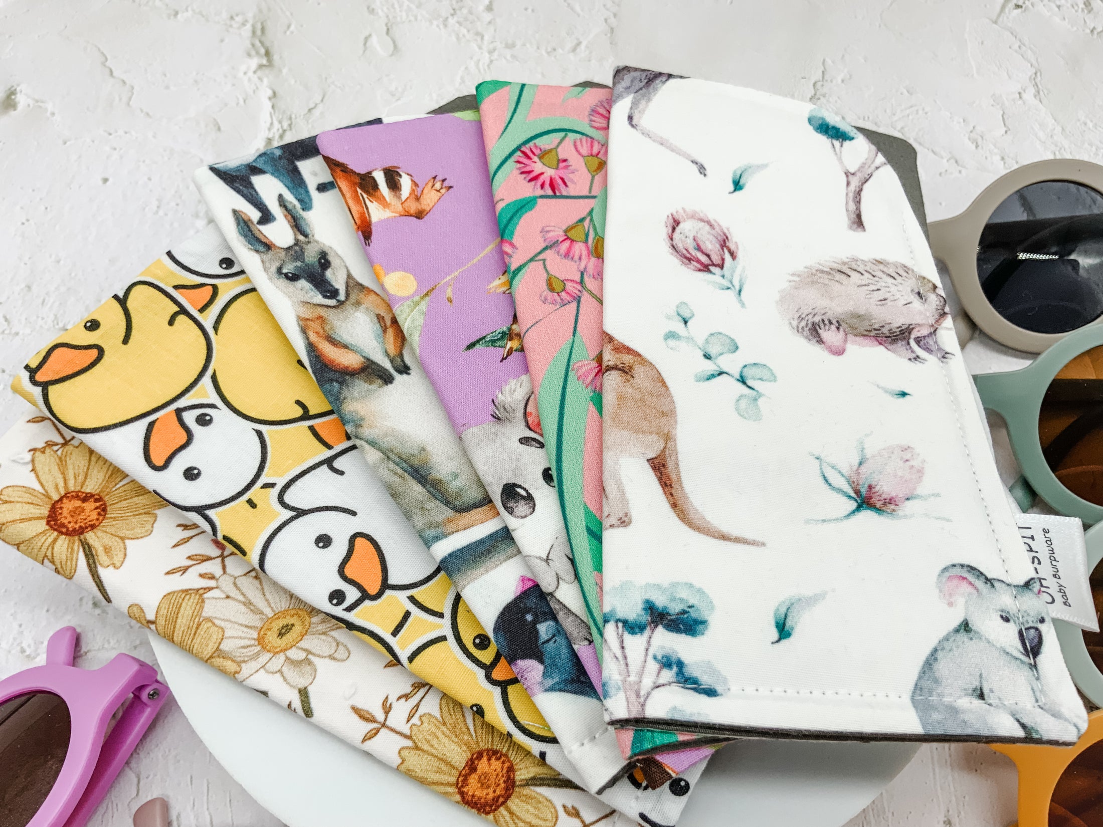 A close-up view of colourful baby bibs featuring playful prints like ducks, flowers, and Australian wildlife, neatly layered on a white surface.