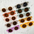 Load image into Gallery viewer, A colourful assortment of baby sunglasses and patterned fabric bibs displayed on a white textured surface, showcasing playful designs and vibrant hues.
