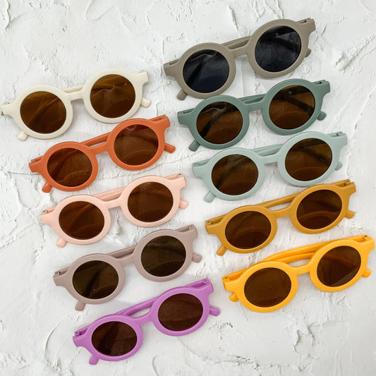 A colourful assortment of baby sunglasses and patterned fabric bibs displayed on a white textured surface, showcasing playful designs and vibrant hues.