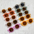 Load image into Gallery viewer, A colourful assortment of baby sunglasses and patterned fabric bibs displayed on a white textured surface, showcasing playful designs and vibrant hues.
