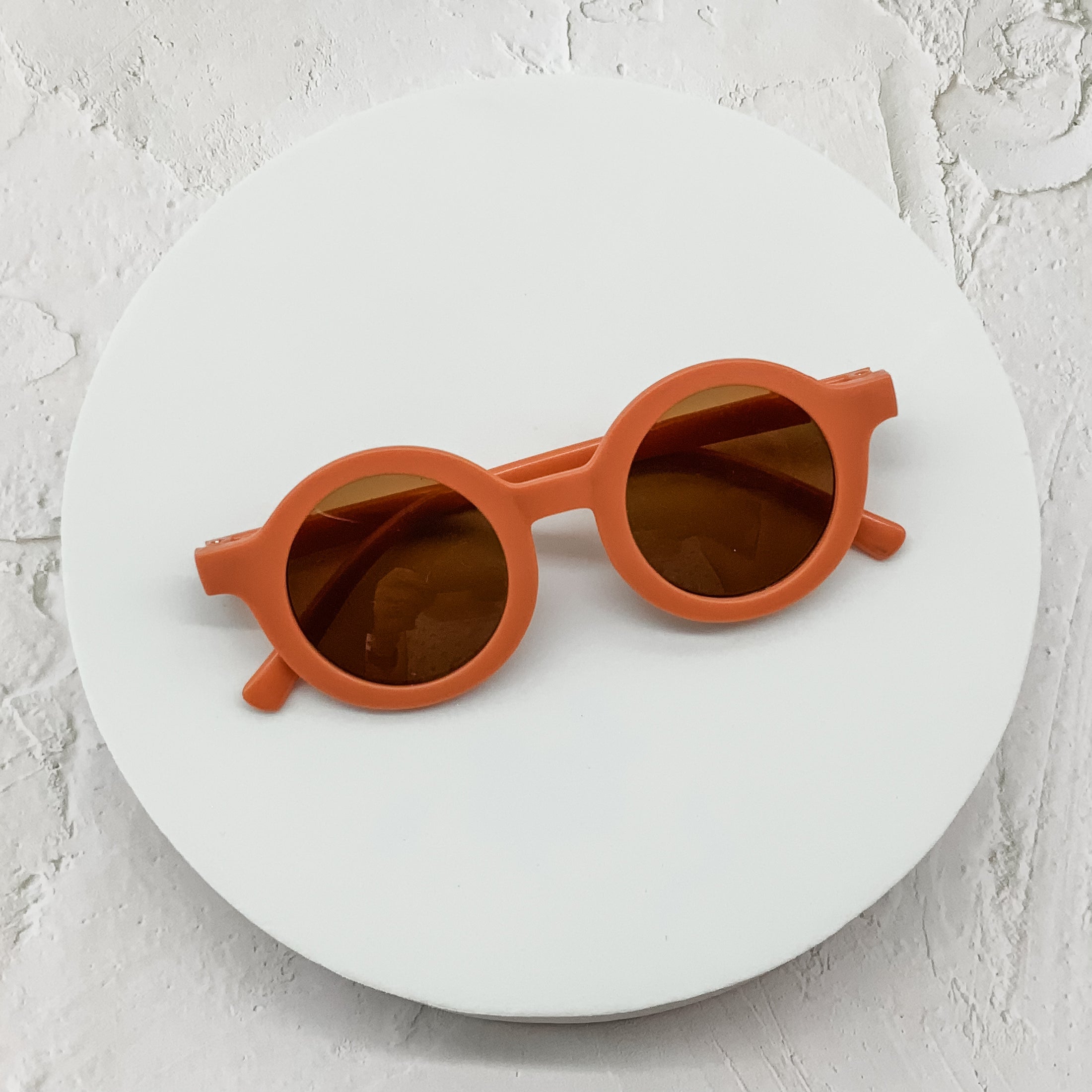 Terracotta orange round sunglasses featuring brown-tinted lenses, elegantly displayed on a white circular backdrop.