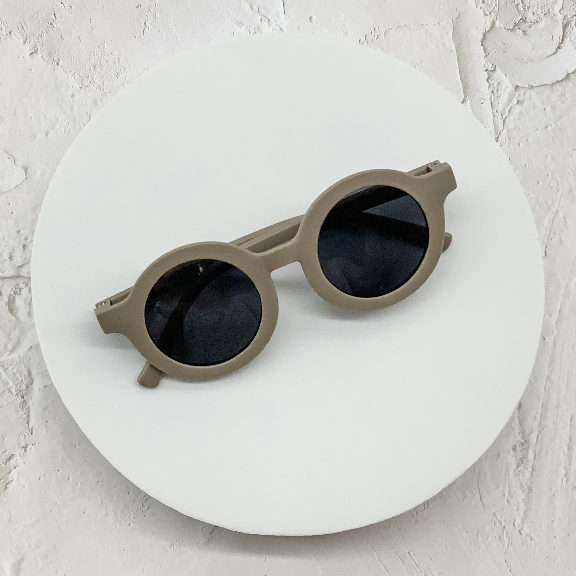 Beige round sunglasses with dark lenses, displayed on a minimalistic white circular platform.
