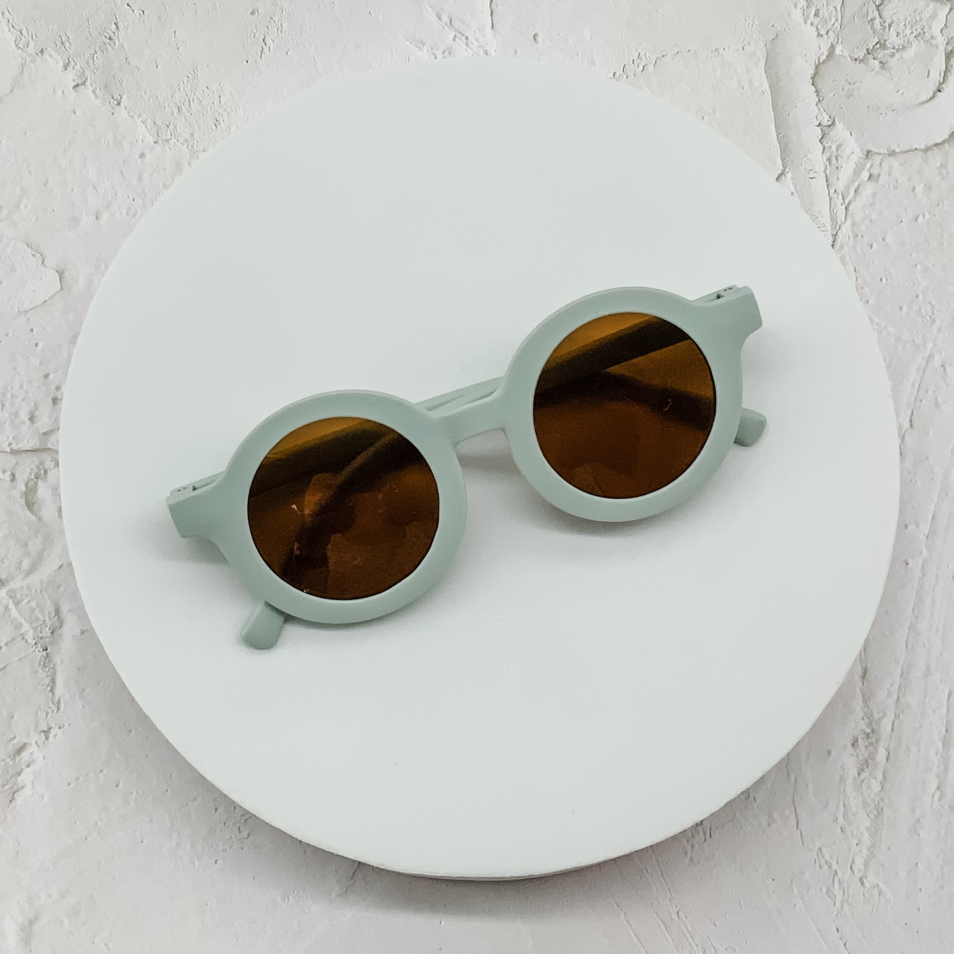 Round pastel green sunglasses with brown lenses, neatly displayed on a minimal white circular surface.