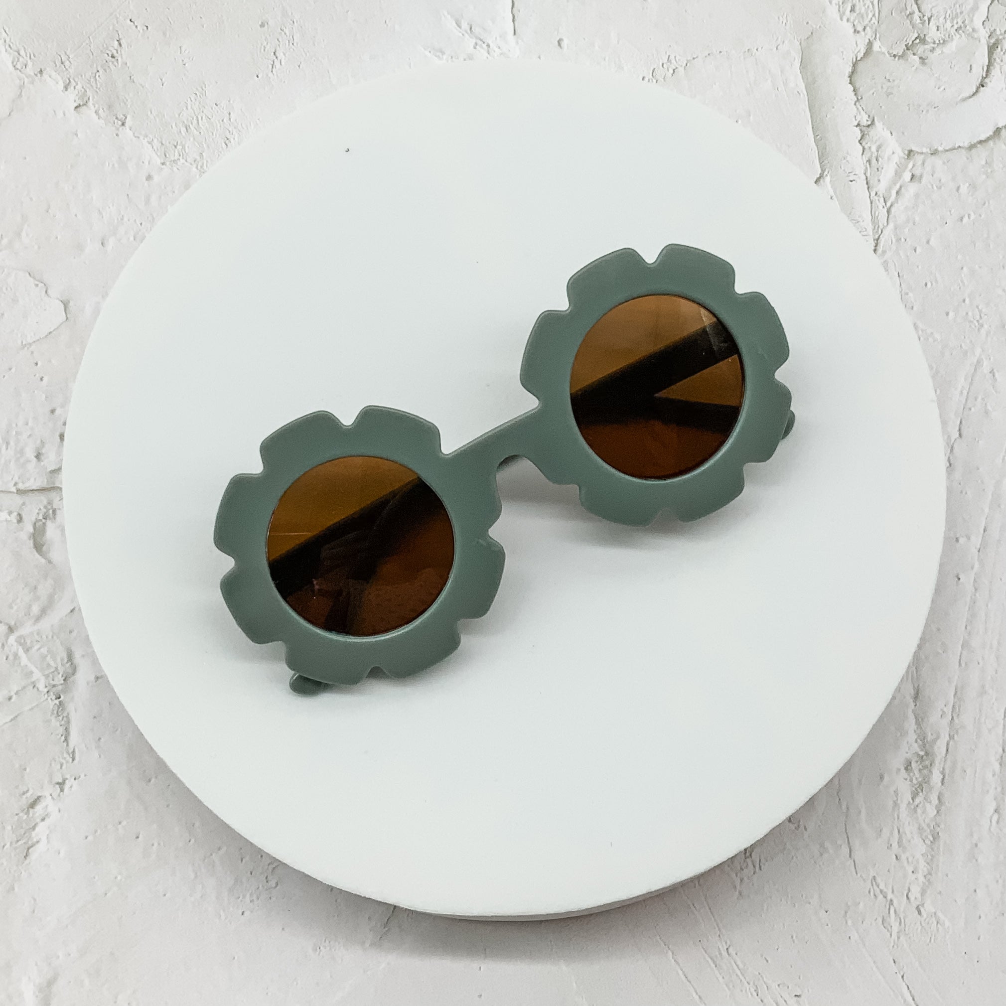 Round sunglasses with sage green flower-shaped frames and brown lenses, displayed on a white circular surface.