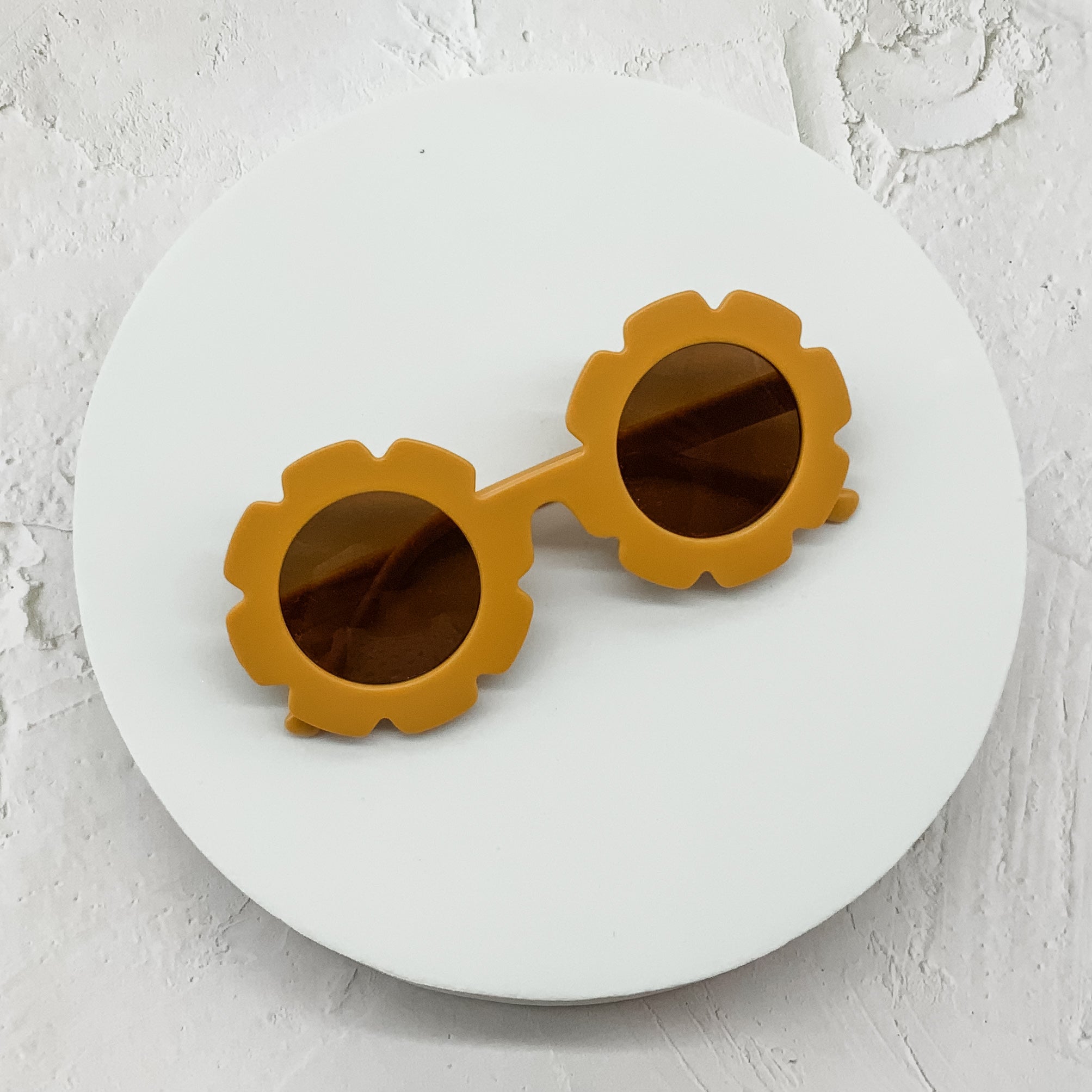 Round sunglasses with mustard yellow flower-shaped frames and brown lenses, displayed on a white circular surface.