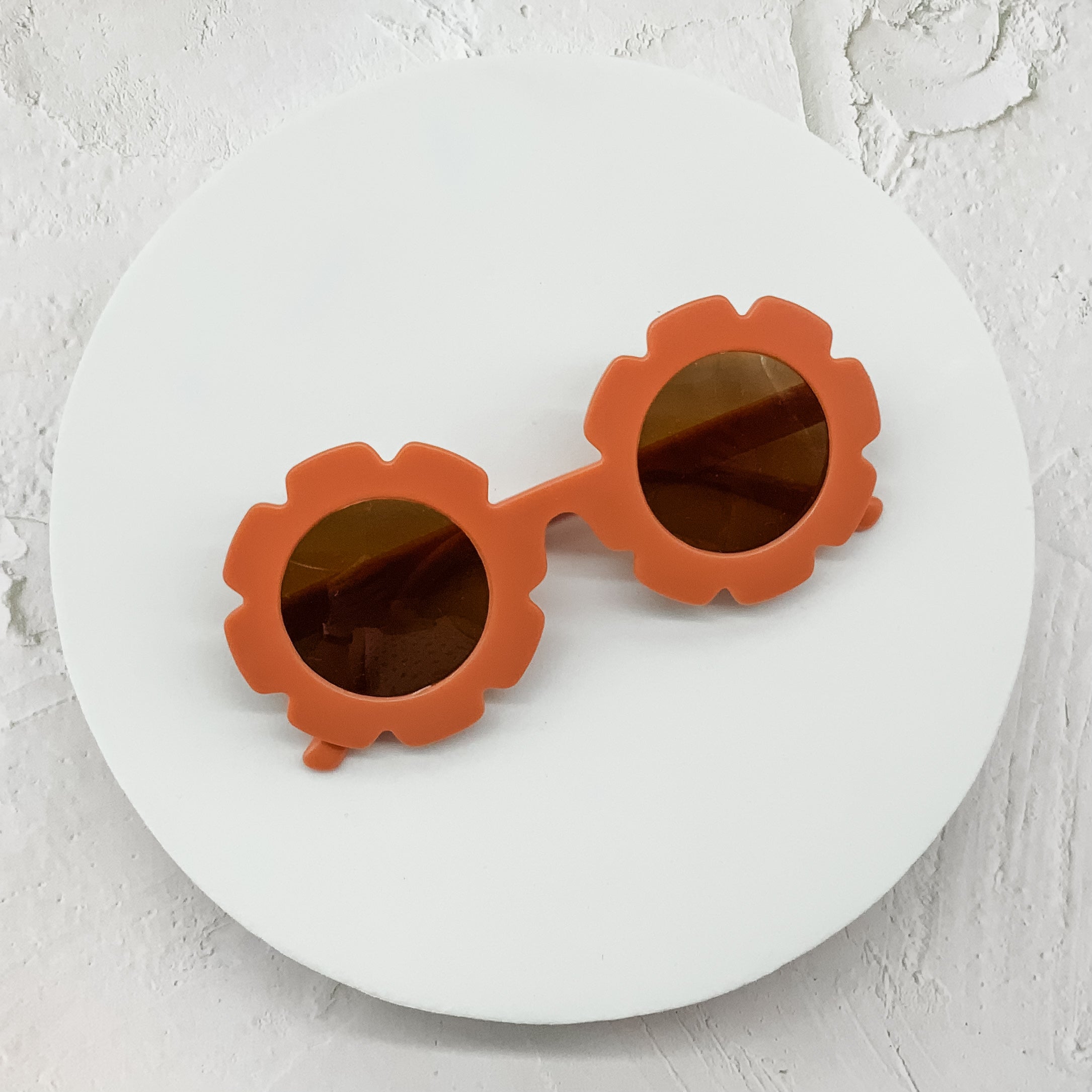 Round sunglasses with rust-coloured flower-shaped frames and brown lenses, displayed on a white circular surface.